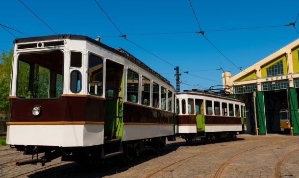 Vintage tram cars parked on cobblestone tracks in a rail yard under a bright blue sky, beside a museum depot with green doors.