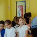 Children participate in a summer camp activity in a yellow classroom; a staff member in a red shirt supervises.