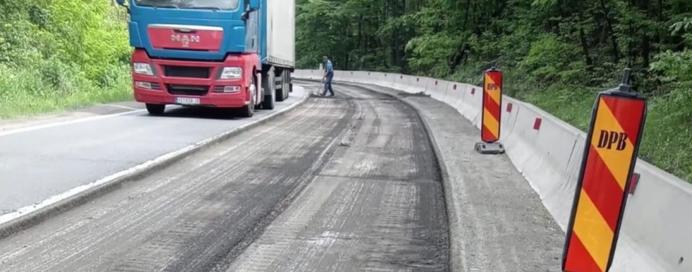 Blue and red semi-truck on a resurfacing road beside wooden barriers in a forested area.