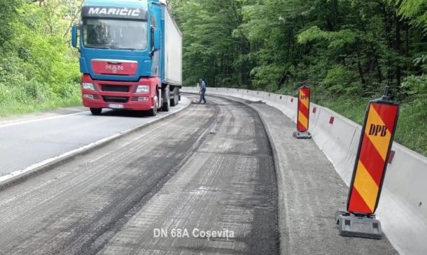 Blue and red semi-truck on a resurfacing road beside wooden barriers in a forested area.