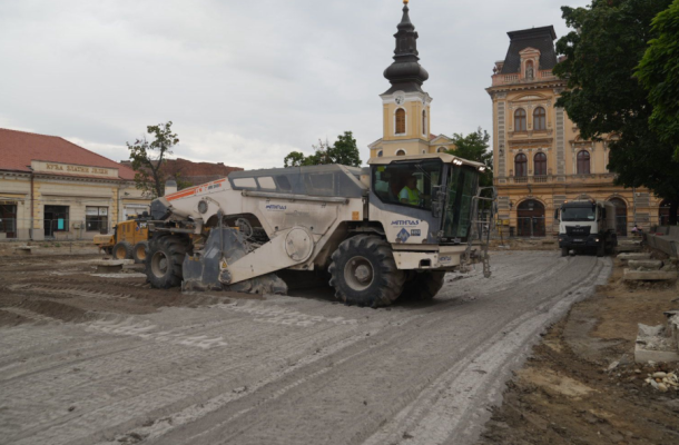 White road roller compactor on a dirt street in a historic town square, with a yellow clock-tower building and old architecture in the background