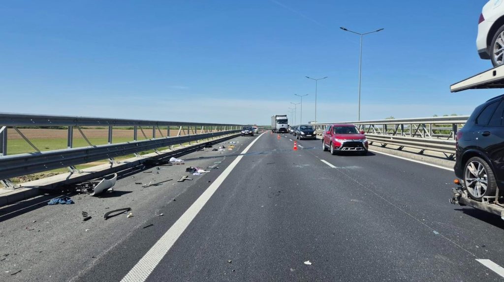 Highway collision scene with debris scattered across lanes and several cars stopped under a clear blue sky.