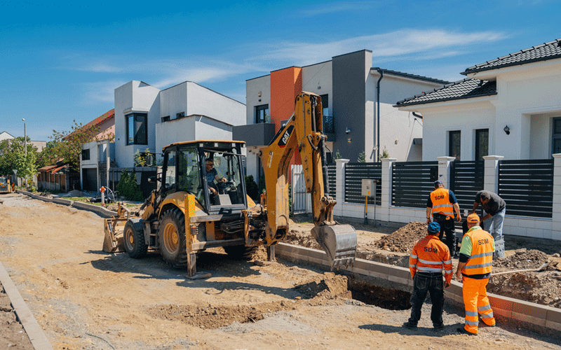 Construction workers in high-visibility clothing supervise an excavator digging a trench in a residential street.