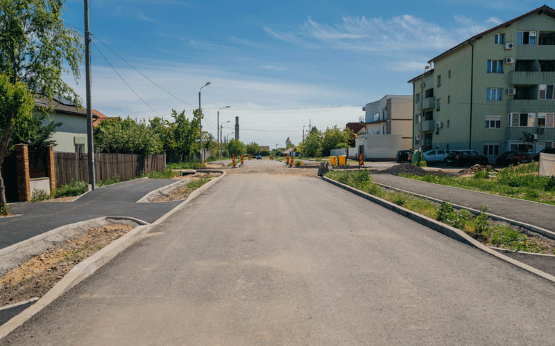 Residential street under construction with orange barriers, sidewalks, and apartment buildings on the right under a blue sky.