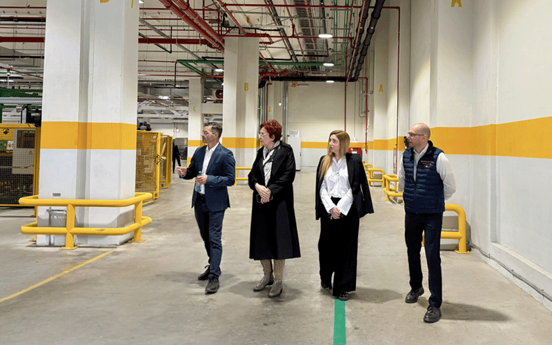 Group of four professionals on a factory tour, walking down a well-lit industrial hallway with yellow safety rails nearby.