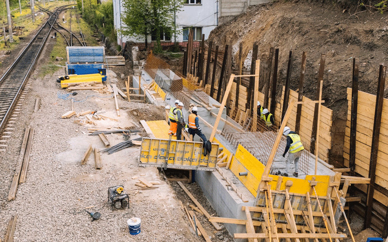 Construction site beside railroad tracks with workers in hard hats and high-visibility vests building a concrete retaining wall, using wooden formwork and steel reinforcement bars.