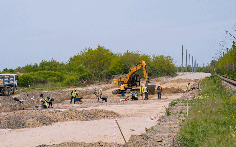 Construction workers in high-visibility vests operate a yellow excavator and shovel dirt beside a railway track as part of a ground-prep project.