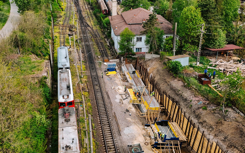 Aerial view of a railway construction site with a passenger train on the left and workers building along the tracks on the right, with houses and trees in the background.