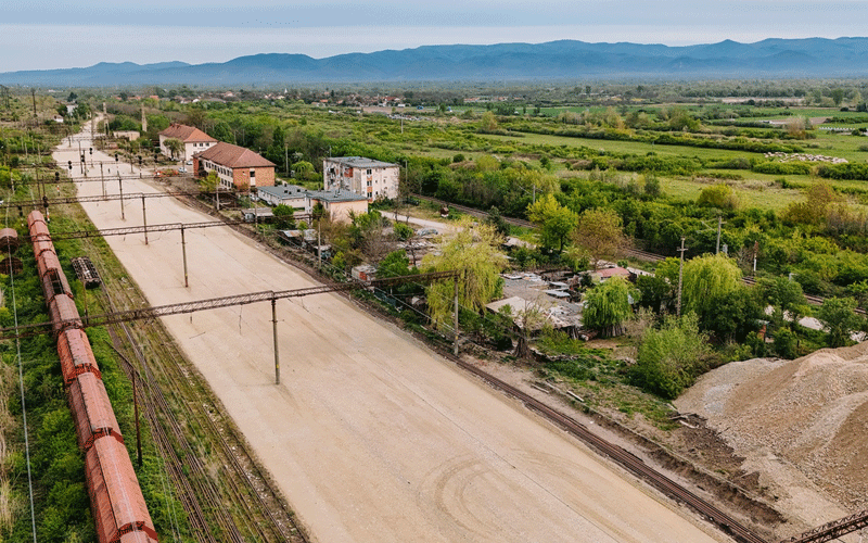 Aerial view of a rural landscape with a long orange freight train on the left, a dirt road, scattered houses, and green fields with distant mountains on the horizon.