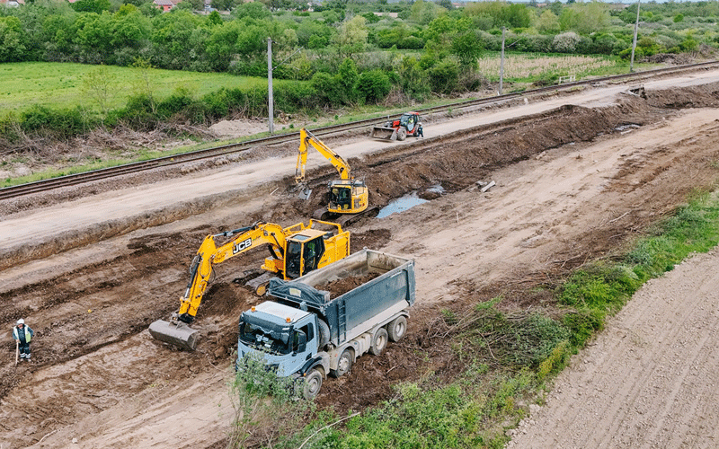 Construction site beside railway tracks with a yellow excavator loading soil into a dump truck; worker nearby.