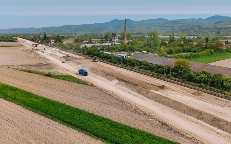 Rural construction site along a dirt road with trucks, near railroad tracks, green fields, and a distant industrial chimney and hills.