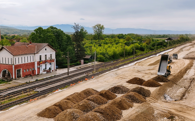 Railway construction site with a dump truck unloading dirt beside tracks and a red brick station building.