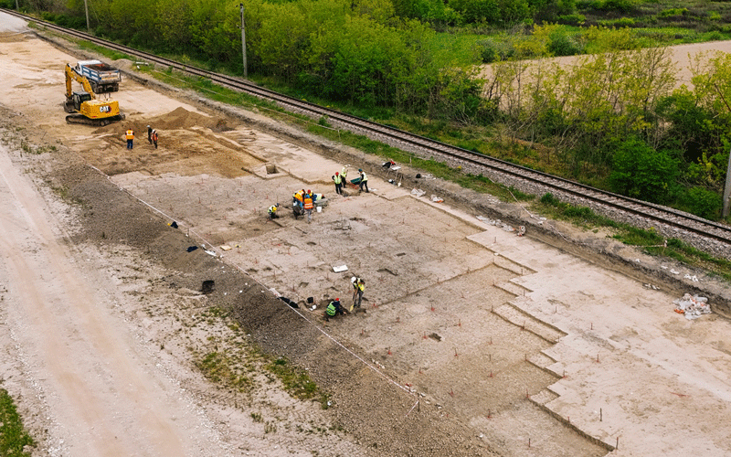Construction crew at a rail-side site with a yellow excavator, grading a dirt area near train tracks from an aerial view.