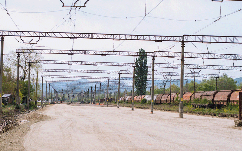 Rail yard with rows of overhead electric wires and tall poles, a dirt road in the center, and rusted freight cars on the right extending toward the horizon.