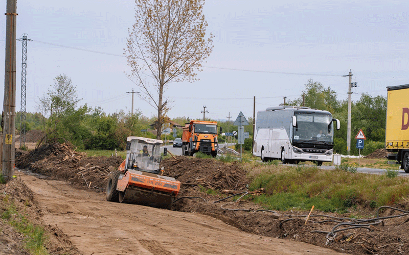 Road construction scene with an orange dirt compactor on a muddy path; a white bus and trucks are on the road behind, with trees and utility poles in the background.