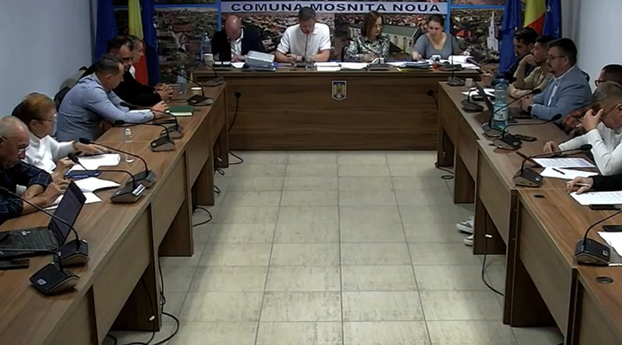 Municipal council meeting in a U-shaped room; officials sit with microphones and papers around long wooden tables, banner reading 'COMUNA MOSNITA NOUA' behind the head table.