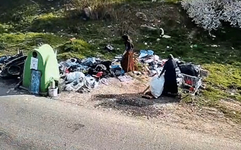 A small green tent beside a roadside trash pile with people sorting through belongings nearby.