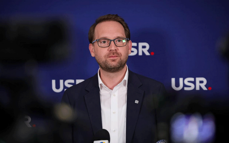 Man in glasses and a dark suit speaks at a press conference with a blue USR backdrop behind him.