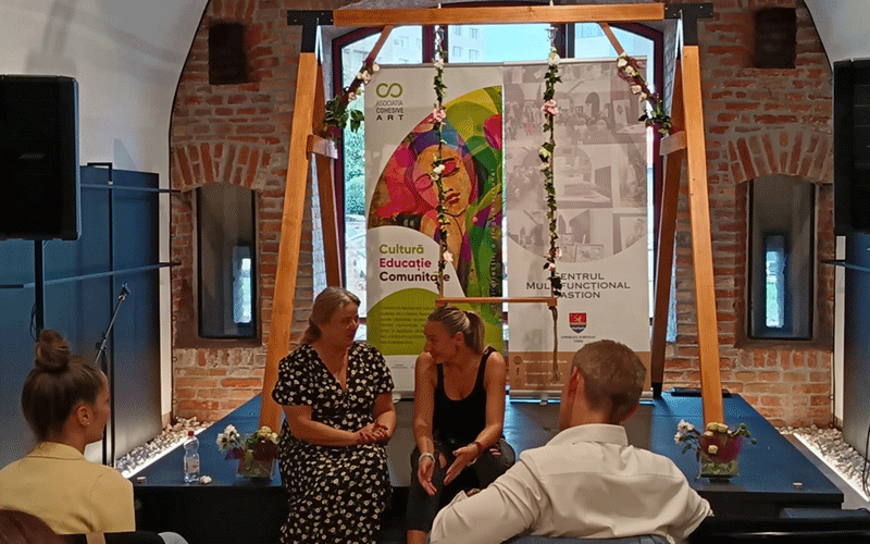 Three people talking and gesturing while seated in front of a wooden arch with banners in a brick-walled room.