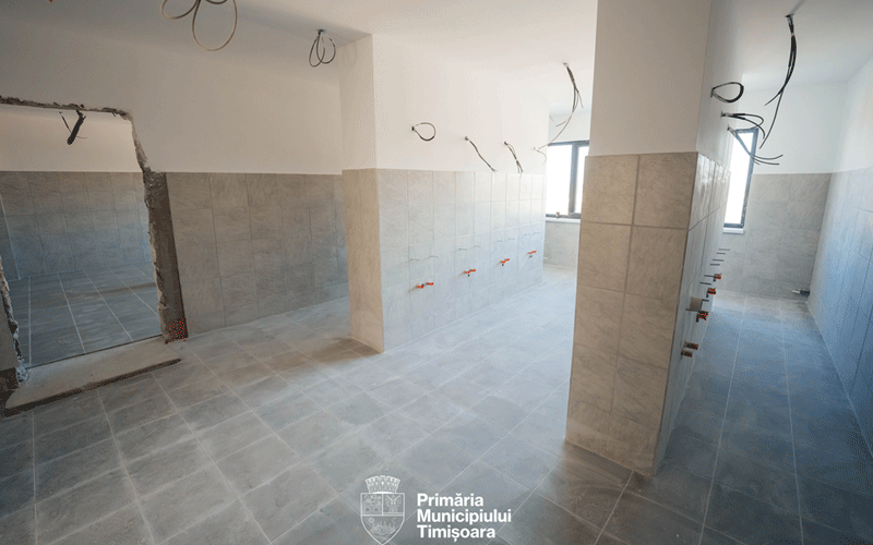 Public restroom under construction in a tiled room with exposed wiring and plumbing rough-ins along the walls.