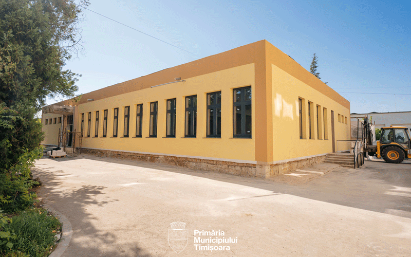 Yellow, single-story municipal building with a long row of tall narrow windows and a construction vehicle on the right side.