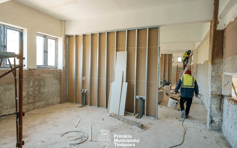 Interior of a building under renovation; workers in hi-vis vests install metal stud walls, with debris on a dusty floor and scaffolding to the left. Logo of Primăria Municipiului Timișoara visible at the bottom.