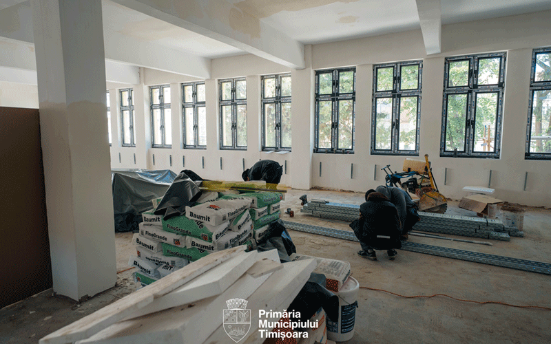 Construction site interior with two workers bending over, stacks of building materials, and metal beams scattered on the floor; large windows along the wall and a city logo at the bottom center.
