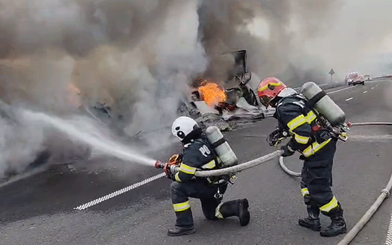 Two firefighters in full turnout gear spray a burning vehicle on a highway while thick smoke erupts and flames glow nearby.