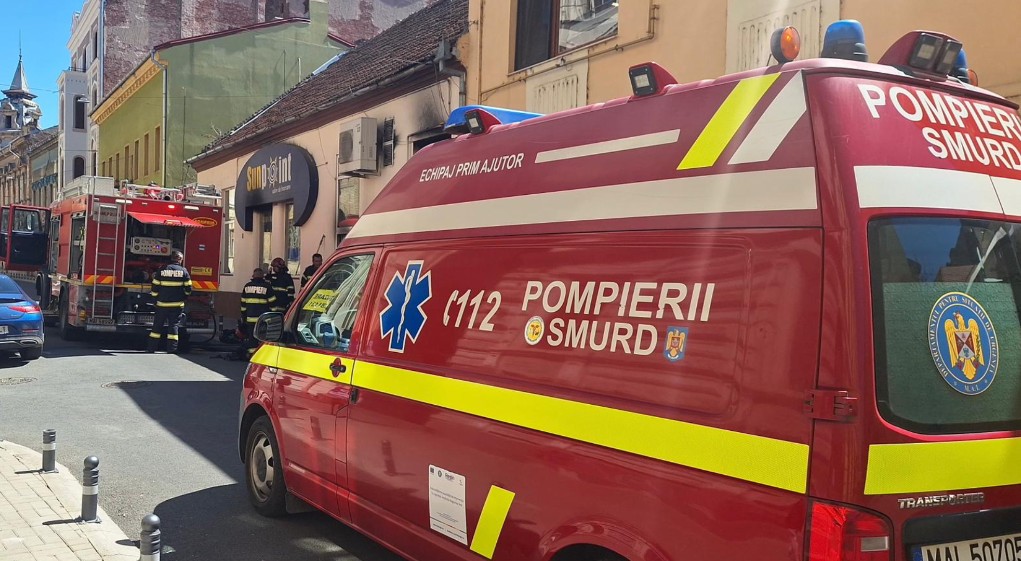 Red SMURD emergency van with yellow reflective stripe parked on a street, firefighters in protective gear in the background