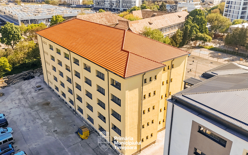 Aerial view of a yellow multi-story building with an orange tiled roof, part of a municipal complex, with surrounding trees and other buildings in the background.