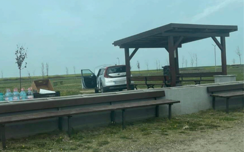 White car with doors open at a park pavilion; water bottles and long picnic benches nearby.