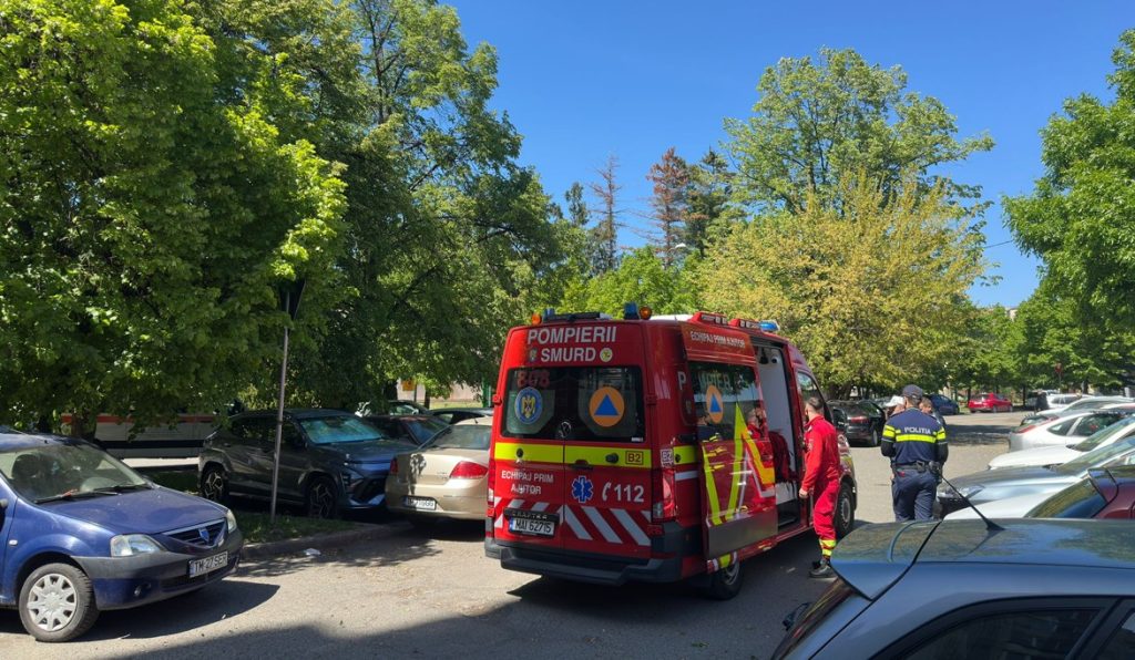 Red emergency ambulance with responders in red uniforms in a tree-lined parking lot under a clear blue sky, preparing for action.