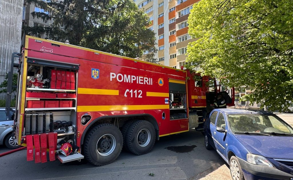 Red fire truck with yellow stripes labeled POMPIERII 112 parked on a city street beside a blue car with a residential building in the background and trees nearby.