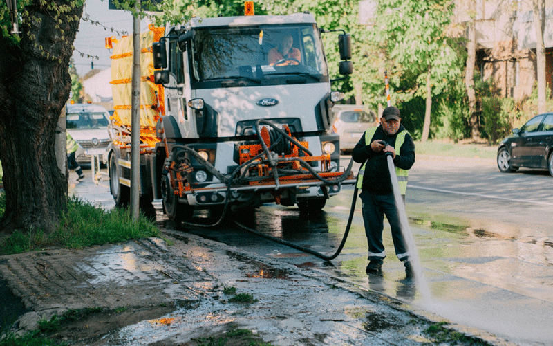 A street crew cleans a puddled road with a large white utility truck, spraying water from a hose held by a worker in a safety vest and cap.