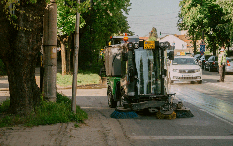 Street sweeper at work on a city street with trees and parked cars along the curb.