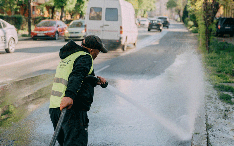 Maintenance worker in a reflective safety vest hosing down a street with a high-pressure nozzle.