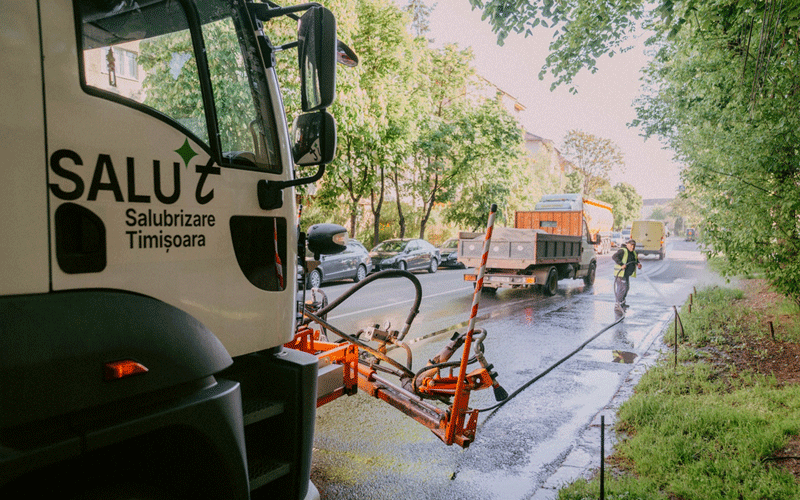 Maintenance truck spraying water on a wet street; worker in a high-visibility vest directs traffic on a tree-lined urban road.