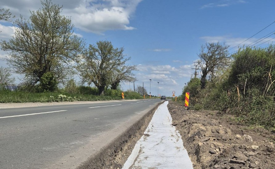 Rural road under construction with a narrow shoulder and a freshly poured white edge line; trees and blue sky above.