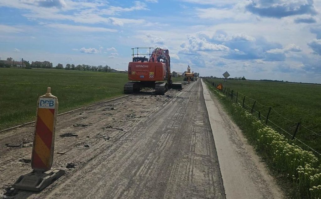 Rural road under repair with an orange Hitachi excavator and a traffic barrier in the foreground.