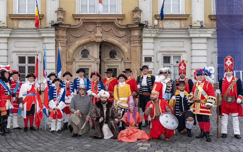 Group of people in colorful historical military uniforms posing for a photo in front of a grand building with an arched doorway on a cobblestone square, likely a reenactment event.