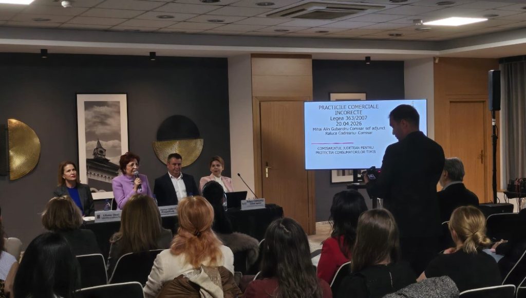 Panel discussion in a conference room: five speakers at a long table, audience seated, and a projector screen displaying a presentation.
