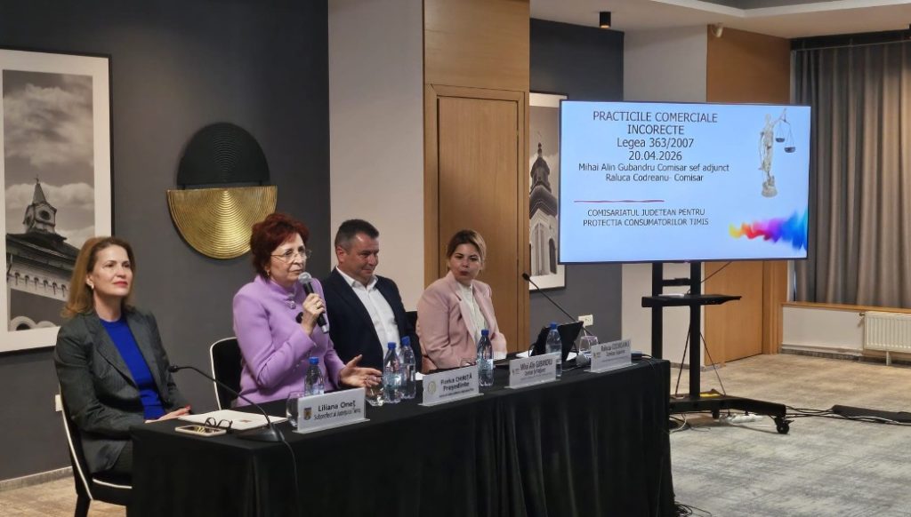 Panel of five professionals seated at a conference table; a woman in a purple blazer speaks into a microphone while others listen. A presentation screen is visible in the background.