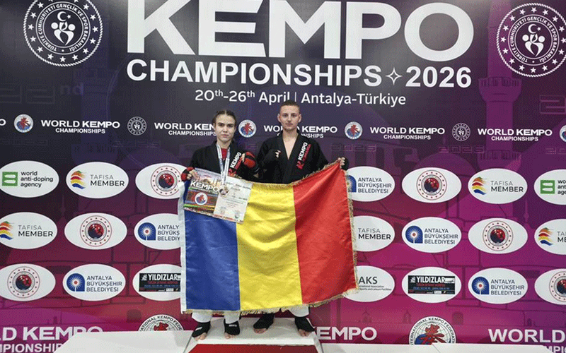 Two athletes stand on a podium at the World Kempo Championships 2026, holding a Romanian flag and certificates, with a backdrop full of logos and the event name.