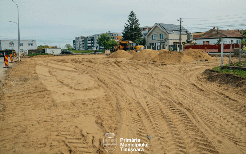 Wide construction site on a residential street with deep tire ruts in sandy soil, piles of sand, heavy machinery in the distance, and houses and utility poles along the road.