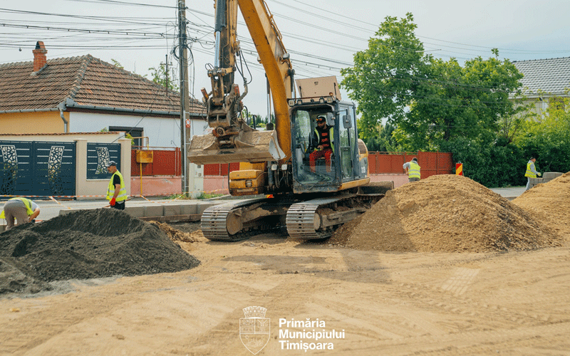 Excavator moving dirt on a street under construction as workers in high-visibility vests level piles of sand and gravel beside residential houses. City logo visible on the ground.
