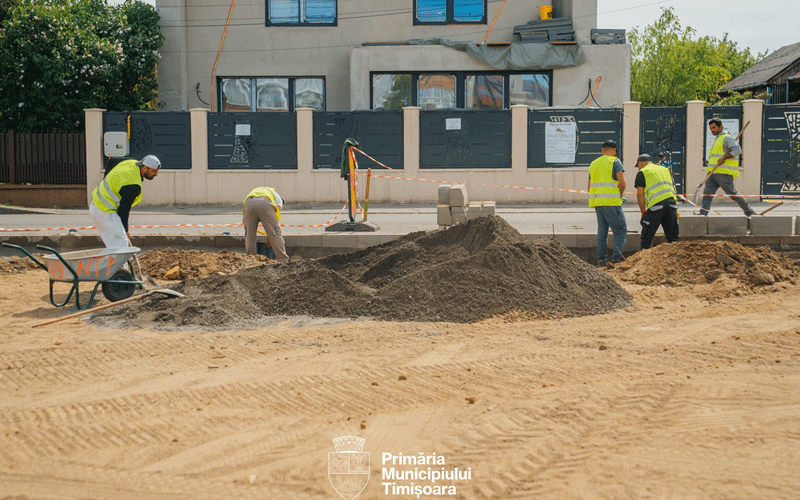 Construction crew in high-visibility vests digging and moving dirt on a street, with piles of soil and a wheelbarrow, and a building under construction in the background.