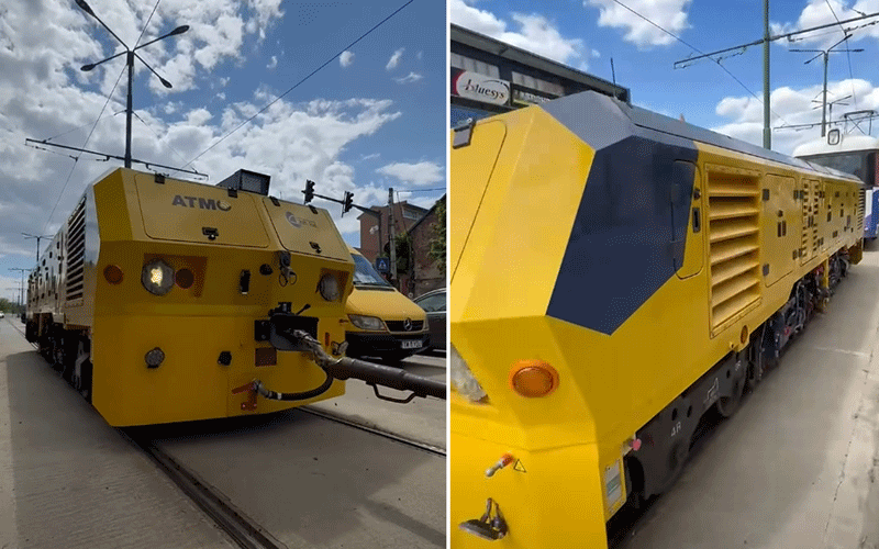 Front view of a large yellow rail maintenance vehicle on tracks with a coupler arm attached to the front.