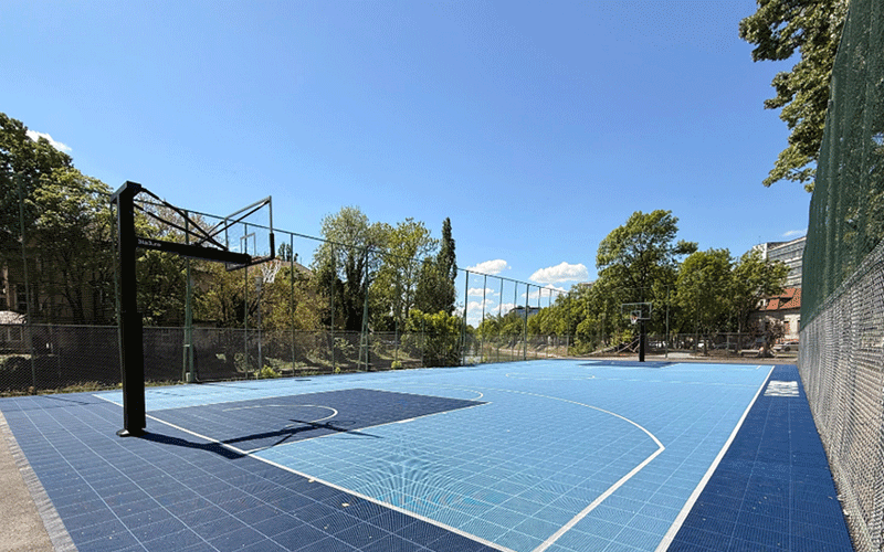 Outdoor blue basketball court with a hoop, surrounded by trees and a chain-link fence under a clear sky