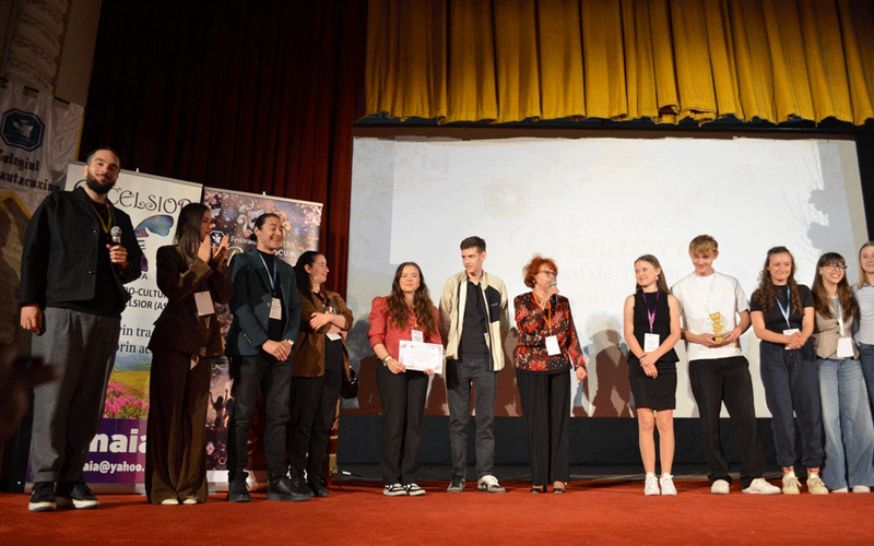 Group of young people on a red-stage, award ceremony vibe; one holds a certificate and another a trophy, with a projection screen in the background.