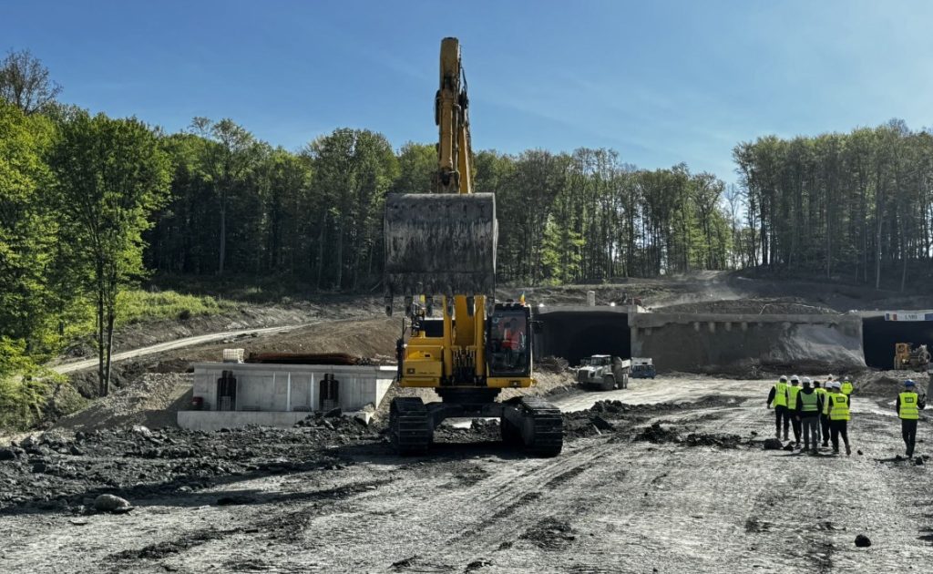 Yellow excavator lifting its bucket at a muddy construction site with a forested backdrop and a tunnel opening nearby. Workers in high‑vis jackets stand to the right, observing.